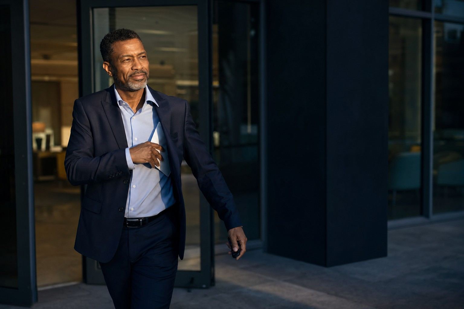 A man in a navy suit walks out of a medical office building in late-afternoon light, tucking a folded sheet of paper into his jacket.